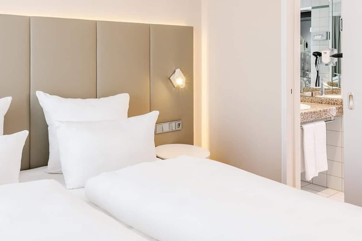 Una habitación de hotel con una cama, una mesita de noche y un baño visible a través de una puerta abierta.