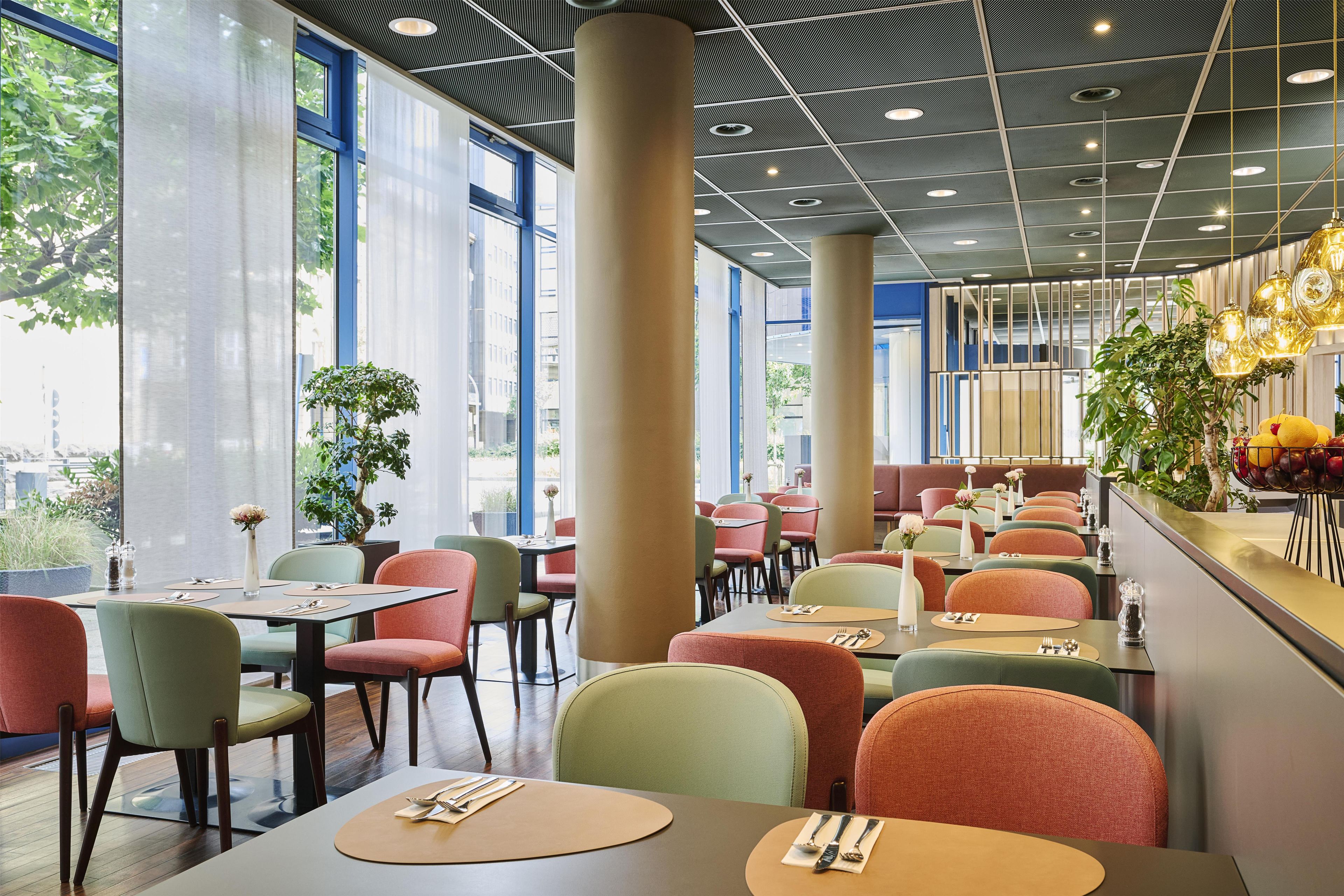 Ein modernes Restaurantambiente mit hohen Decken, großen Fenstern und farbenfroher Sitzgelegenheiten.