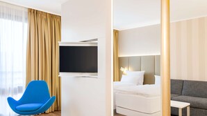 A modern hotel room with a flat-screen TV, a blue chair, a bed, a grey sofa, and a small table. - NH Dortmund (Dortmund)