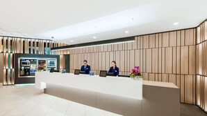 A modern reception desk with two staff members, a vending machine, and a coffee station in the background. - NH Dortmund (Dortmund)