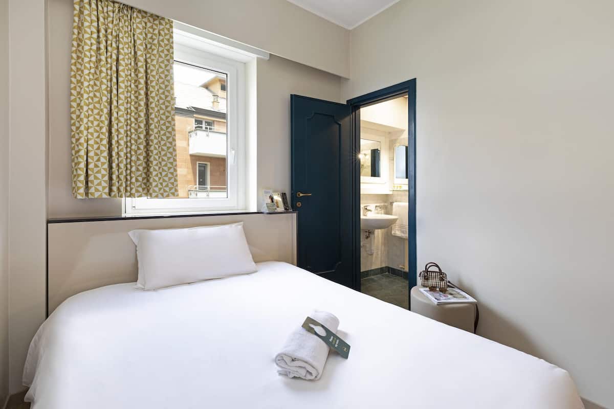 Ein Hotelzimmer mit Bett, einem fenster mit Vorhängen, einer Tür zu einem Badezimmer und einem kleinen Tisch mit einer Zeitschrift und einer Handtasche.