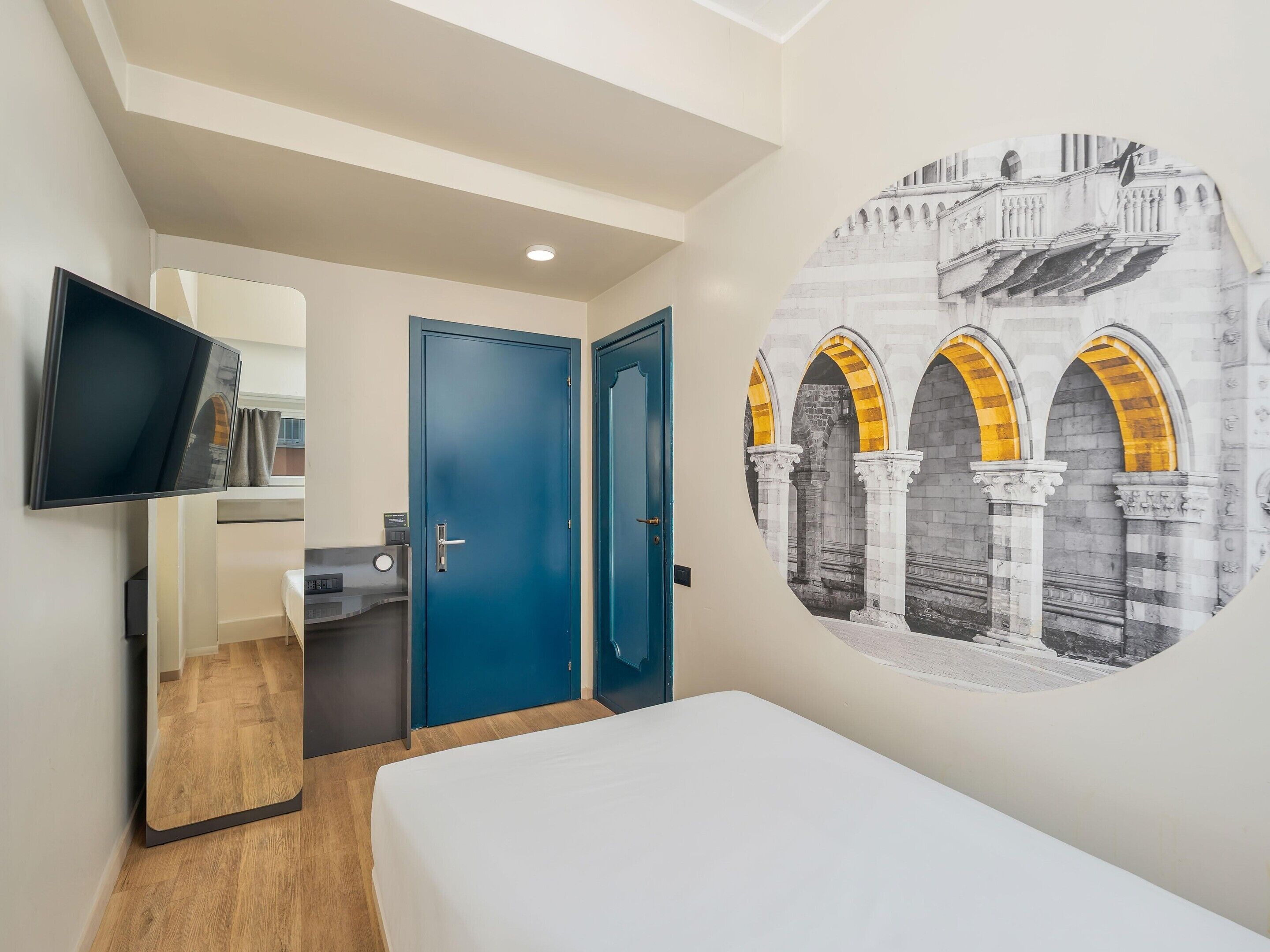 Habitación de hotel con una cama, un televisor fijado a la pared, una puerta y un mural arquitectónico.