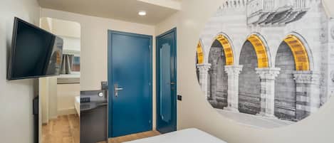 Une chambre d’hôtel comprenant un lit, un téléviseur fixé au mur, une porte et une fresque murale représentant une structure architecturale.