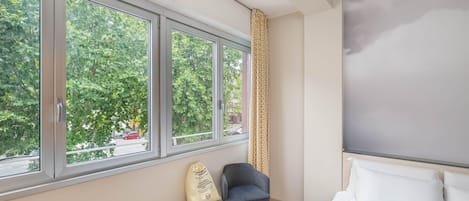 A hotel room with a bed, a chair, a small stool, and a window with a view of trees and a street.