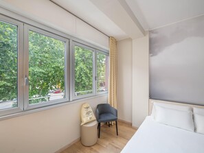 A hotel room with a bed, a chair, a small stool, and a window with a view of trees and a street. - B&B Hotel Como City Center (Como)