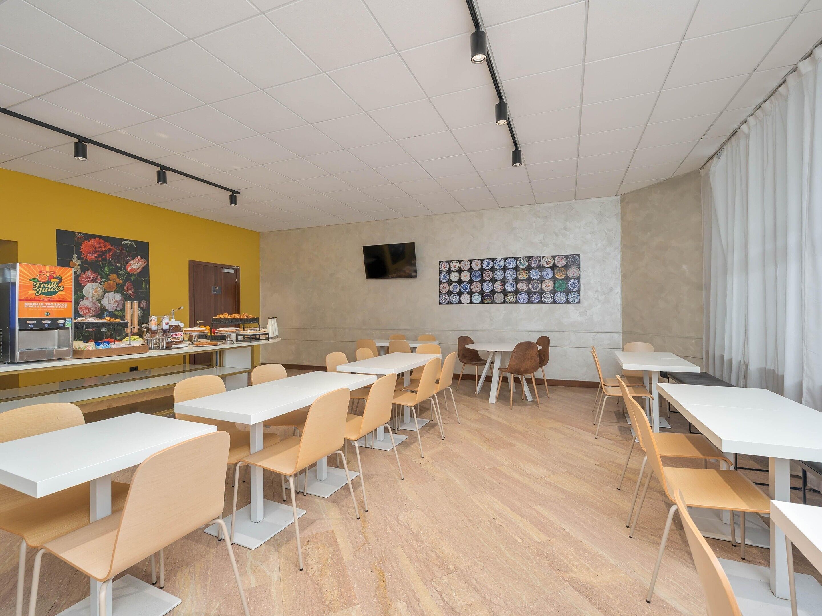 Una cafetería con mesas y sillas blancas, un dispensador de zumo de frutas y un televisor montado en la pared.