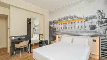 Ein Hotelzimmer mit Bett, Schreibtisch, Stuhl, Spiegel und einem an der Wand befestigten Kunstwerk.