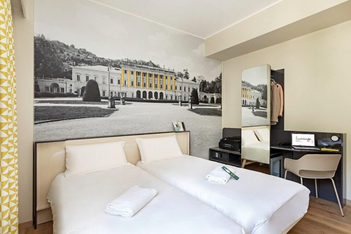 Ein Hotelzimmer mit einem großen Bett, einem Schreibtisch und einem Stuhl. Es gibt ein großes Fenster mit Blick auf ein Gebäude und Bäume.