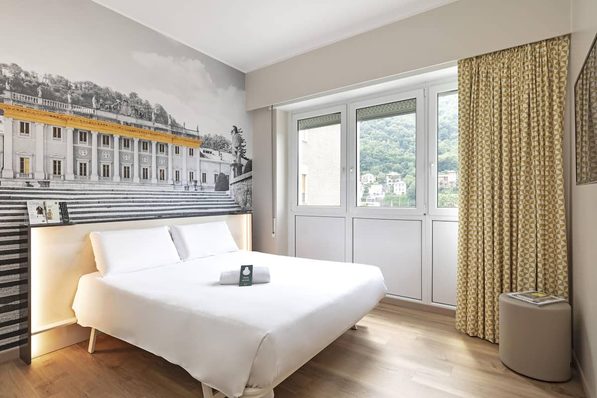 Ein Hotelzimmer mit Bett, einem großen Fenster mit Vorhängen, einem Wandgemälde und einer kleinen Mülleimer.