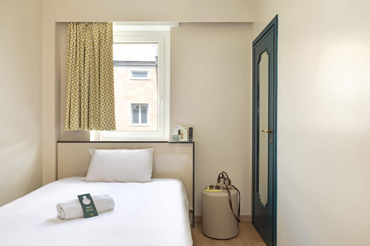 Ein Hotelzimmer mit ordentlich bezogenem Bett, einem Fenster mit gemusterten Vorhängen, einem Nachttisch mit Fernbedienung, einem kleinen runden Hocker und einer Tür mit dekorativem Spiegel.