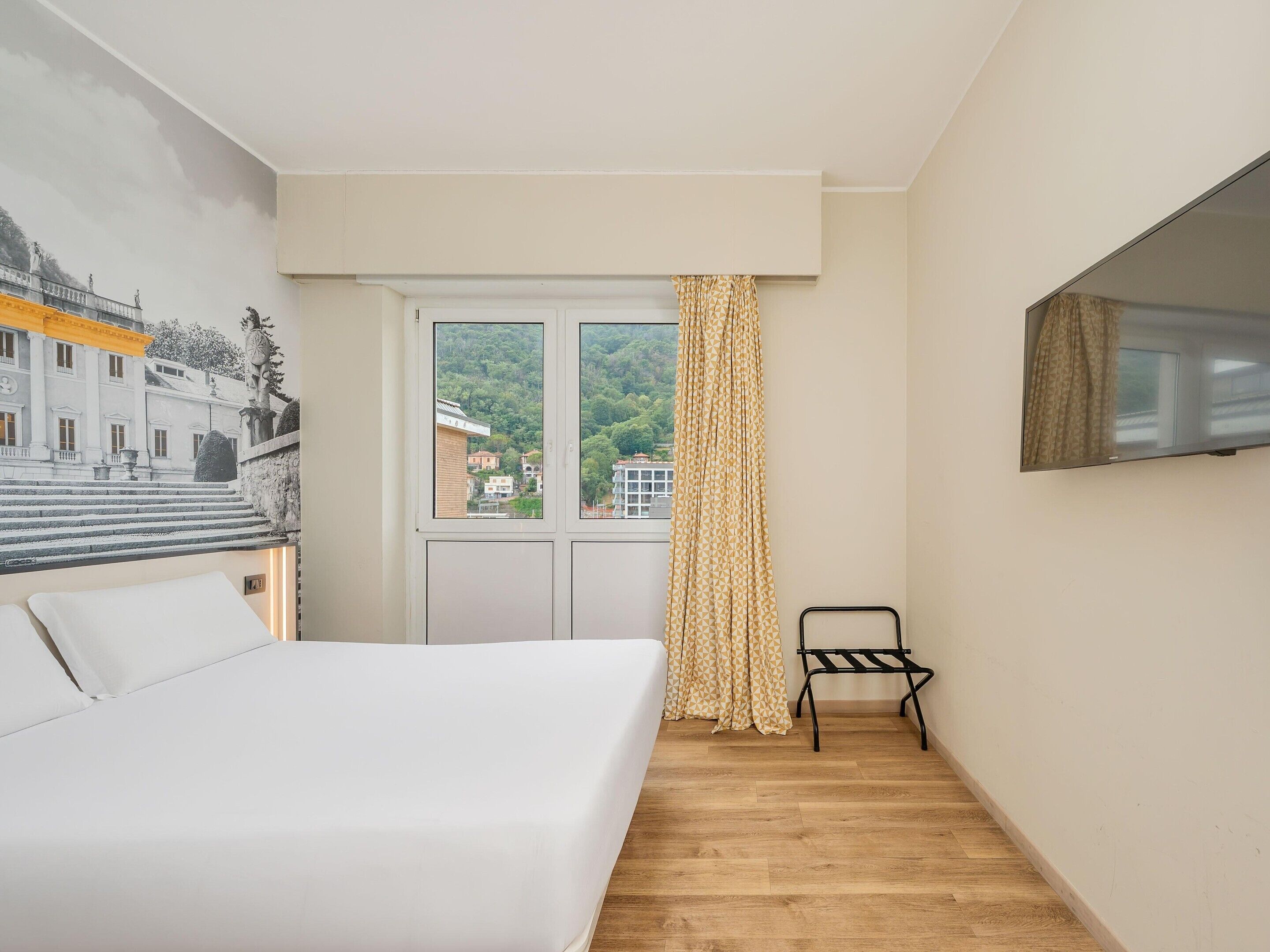 A bedroom with a bed, a window with curtains, a chair, and a wall-mounted TV.