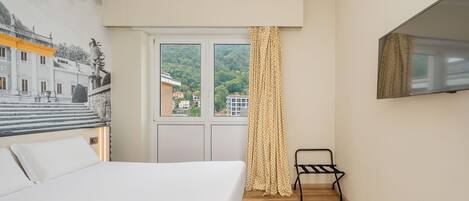 A bedroom with a bed, a window with curtains, a chair, and a wall-mounted TV.