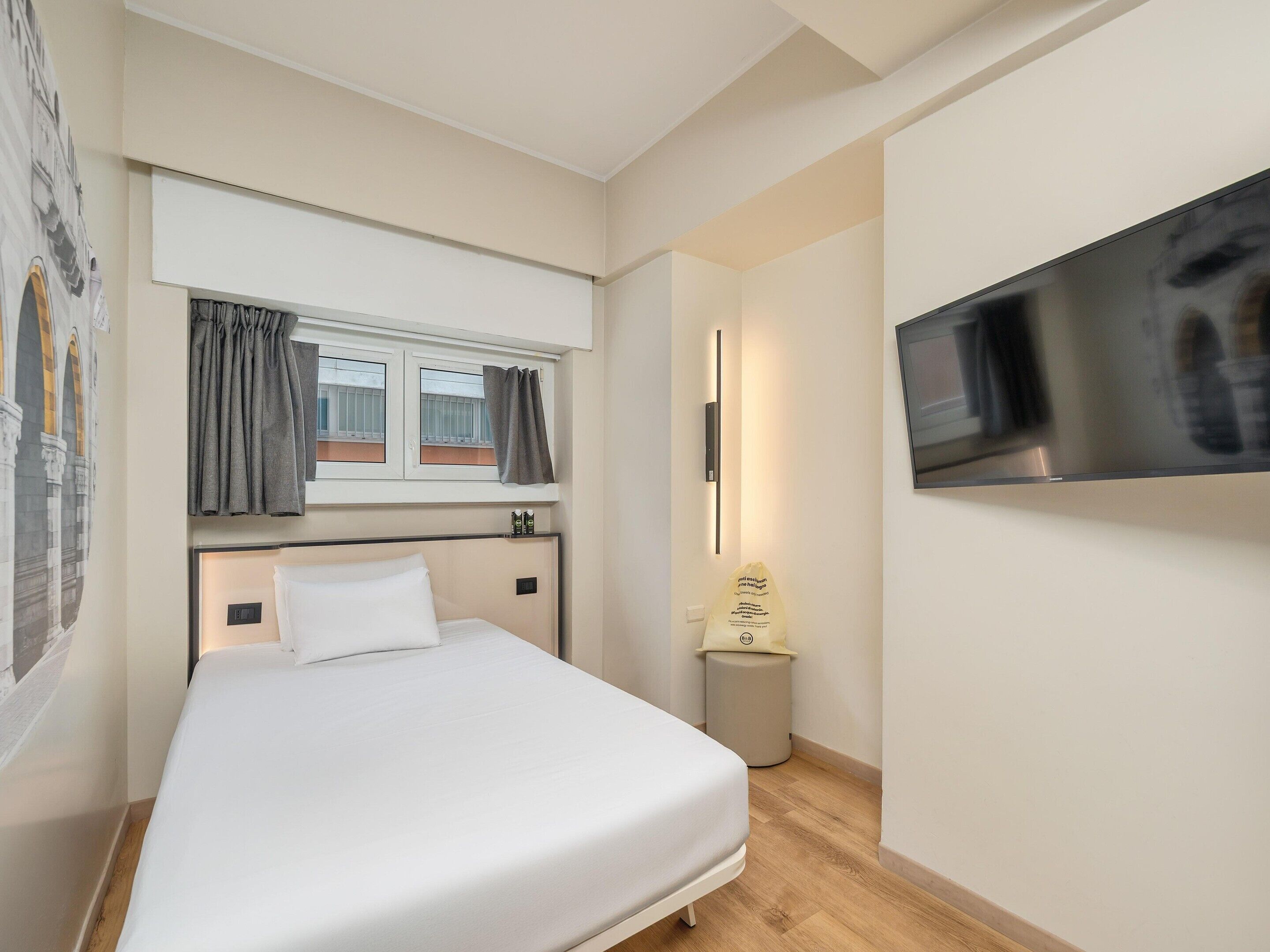 A hotel room with a bed, a window with curtains, a wall-mounted TV, and a small trash bin.