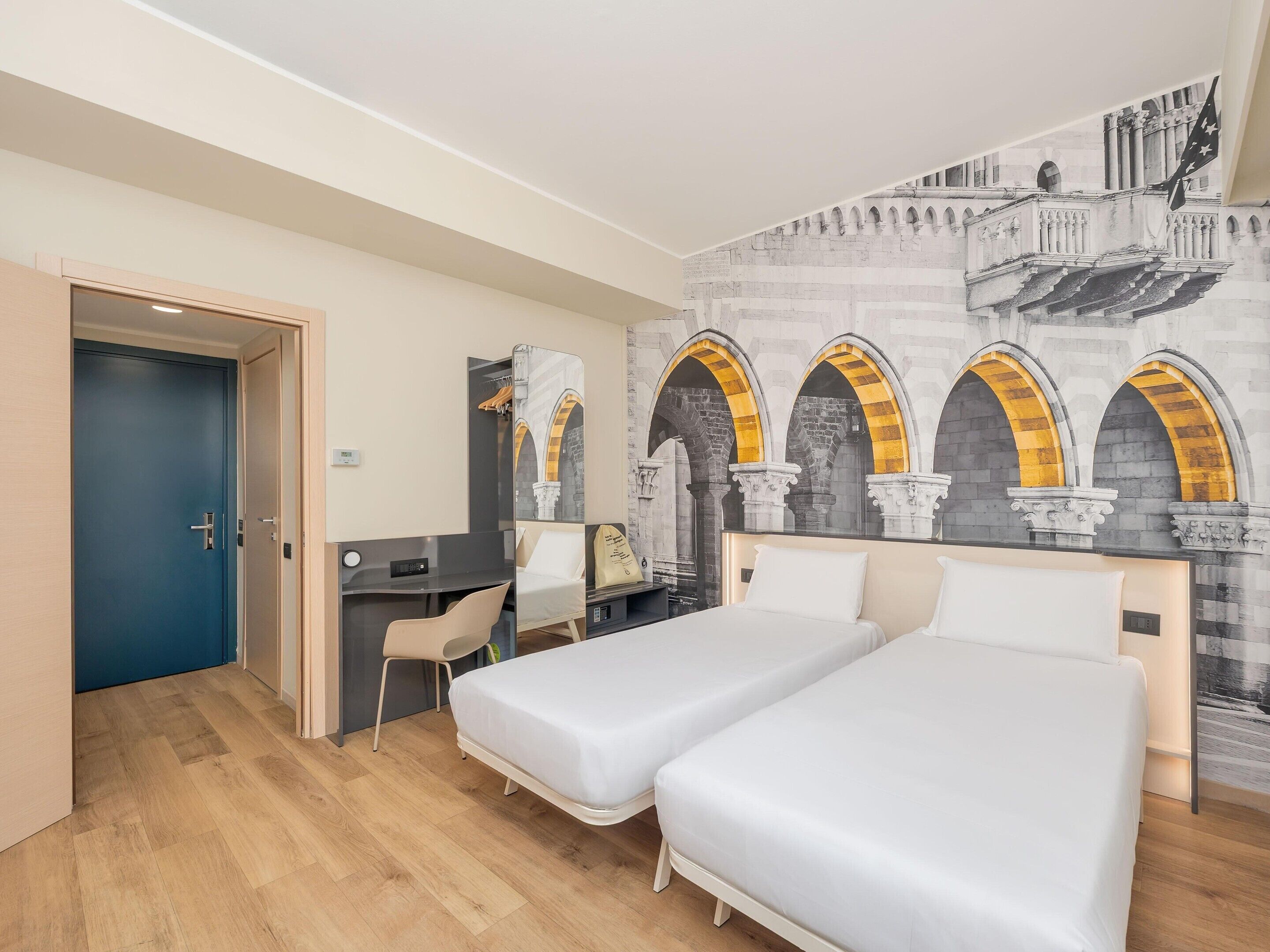 Habitació d'hotel amb dos llits, una taula de treball, una cadira i un mural d'arcs.
