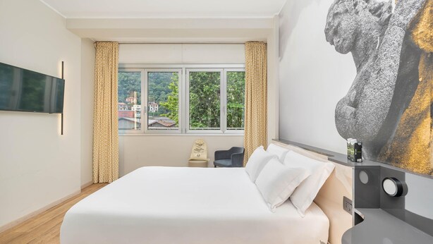 Habitación de hotel moderna con una cama grande, un televisor de pantalla plana en la pared, una ventana con cortinas y un cuadro de una cabeza de león en la pared.