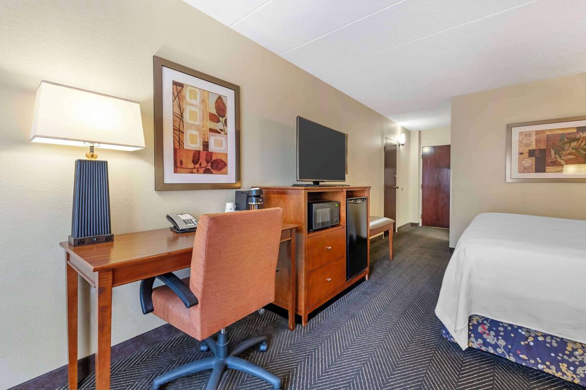 A hotel room with a wooden desk, a chair, a television, a minibar, and a bed.