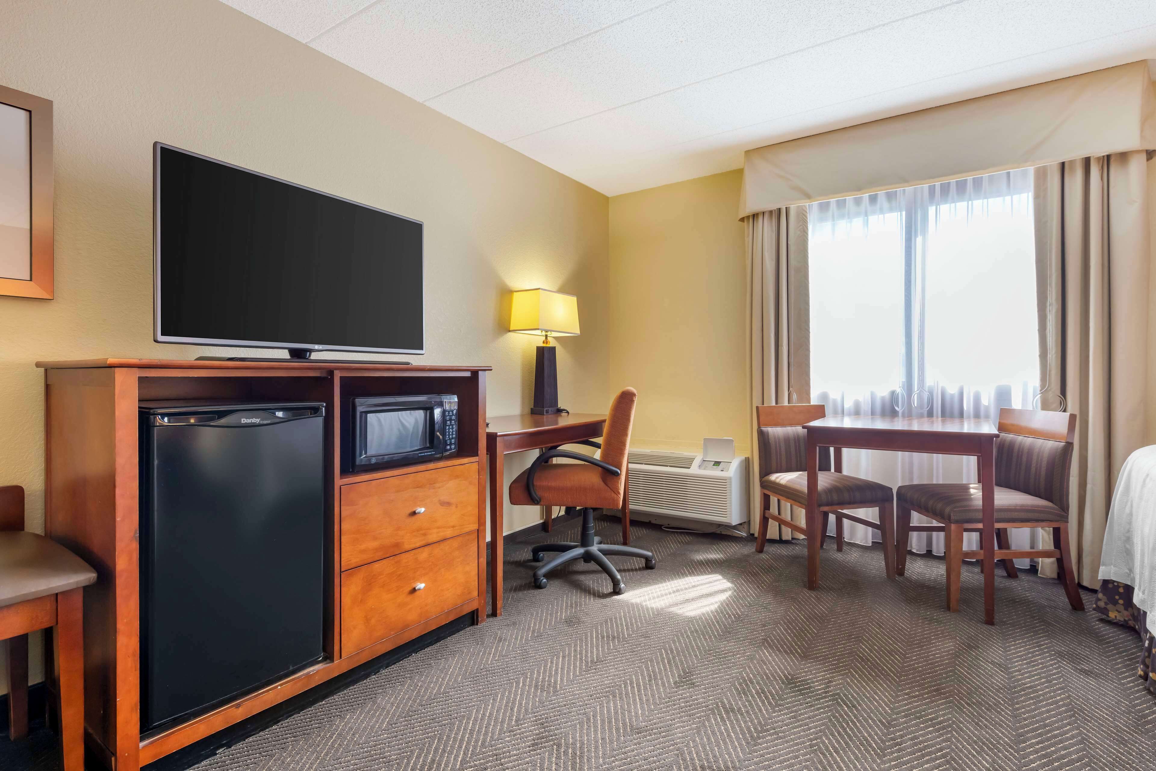 A hotel room with a flat-screen TV, a mini-fridge, a desk with a chair, a dining table with four chairs, and a large window with curtains.