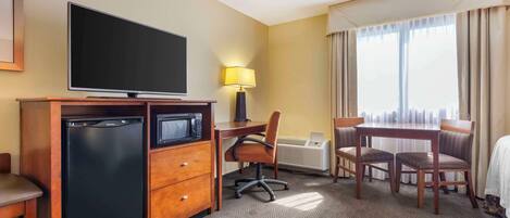 A hotel room with a flat-screen TV, a mini-fridge, a desk with a chair, a dining table with four chairs, and a large window with curtains.