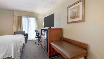 A hotel room with a bed, a desk with a computer, a television, a chair, and a bench.