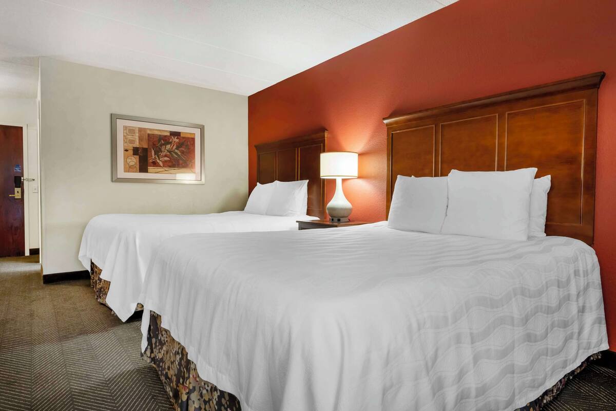 A hotel room with two beds, a nightstand, a lamp, and a painting on the wall.