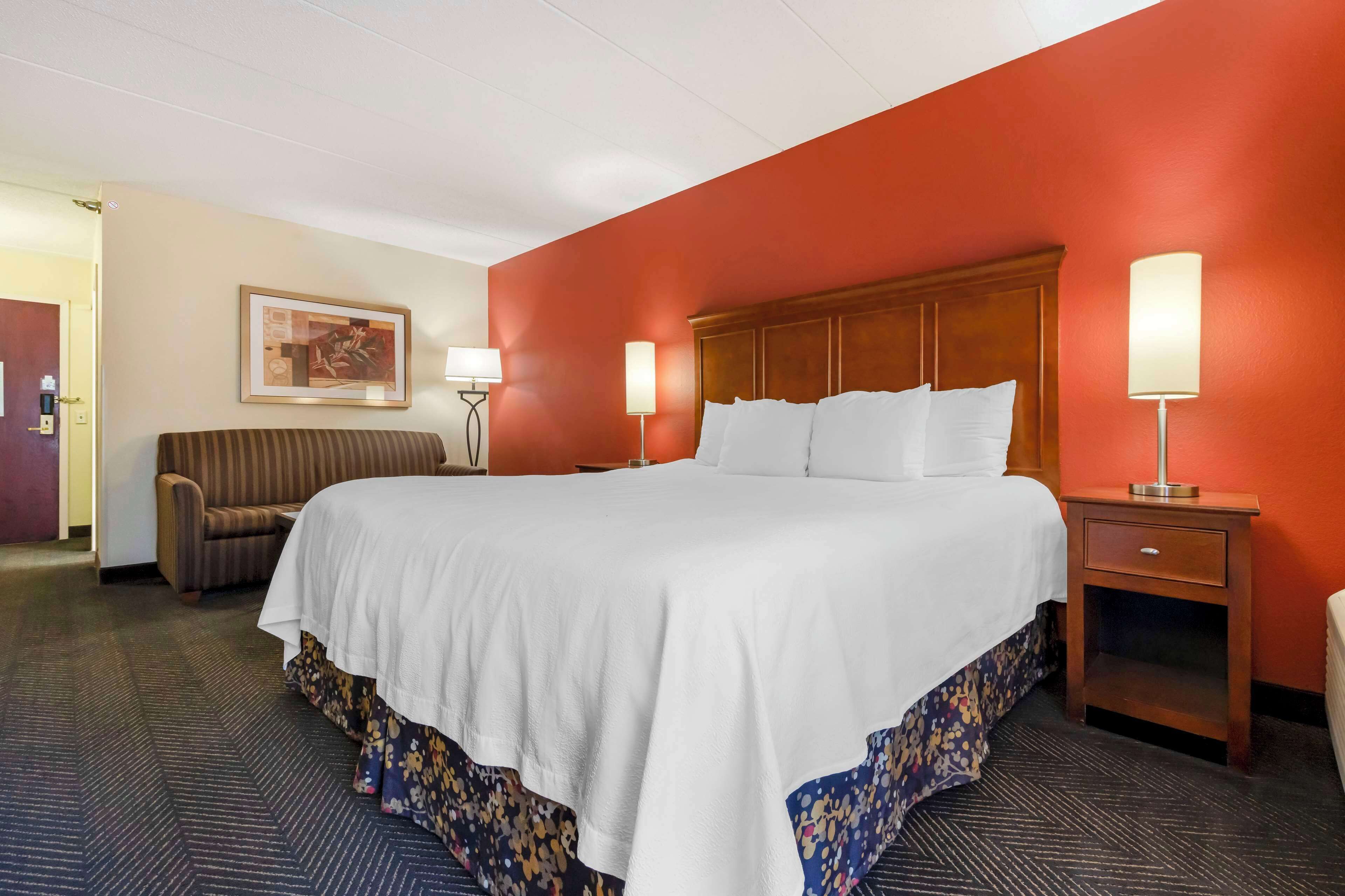 Habitación de hotel con una cama grande, cabecera de madera, dos lámparas de noche, una mesita de noche, un sofá y un cuadro en la pared.