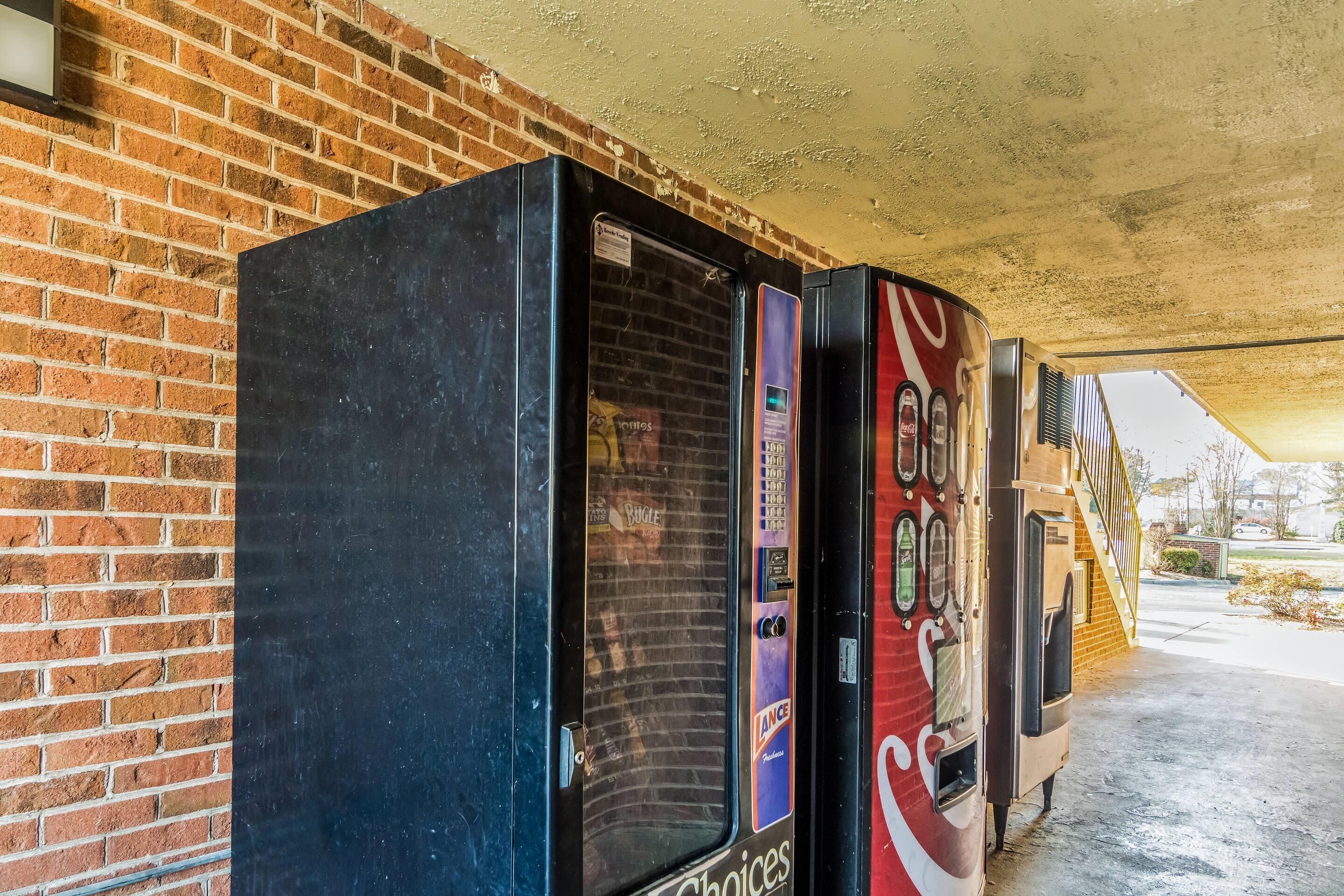 Vending machines with beverage options, a brick wall, and a covered area with a staircase.