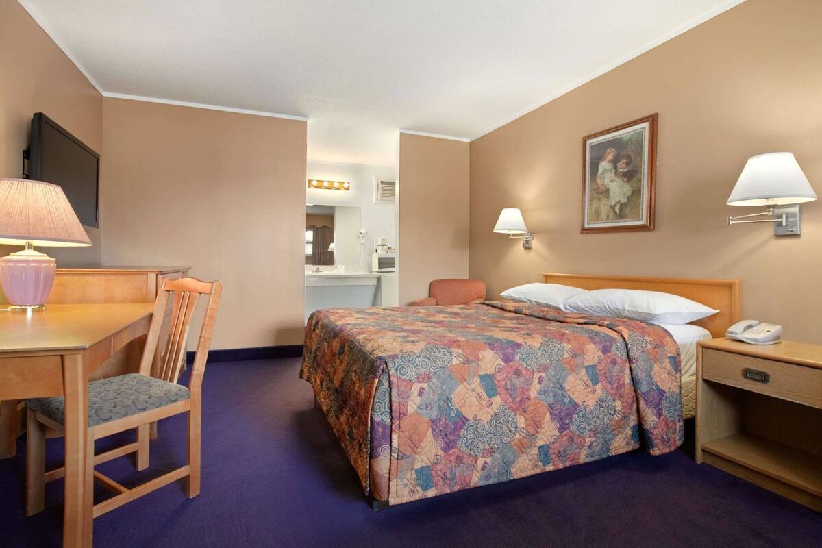 Habitación de hotel con cama, escritorio con lámpara, silla, mesita de noche y un cuadro en la pared.