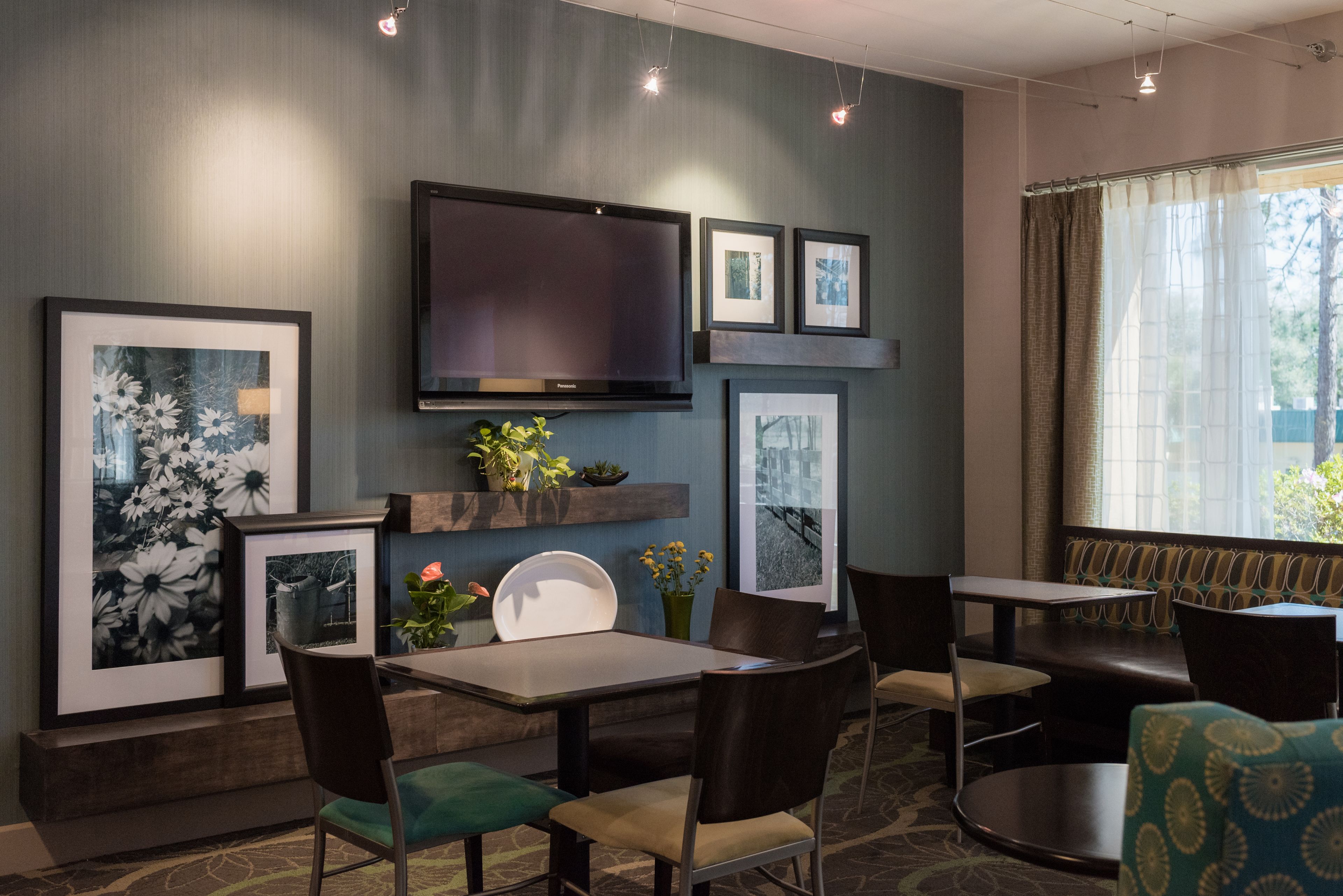 Une chambre d’hôtel moderne dotée d’un téléviseur à écran plat, d’œuvres d’art encadrées, d’une petite plante et d’un coin salon avec des fauteuils et une table.