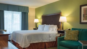 Una habitación de hotel con una cama, dos lámparas de noche, un sillón verde, una mesita y una ventana con cortinas.