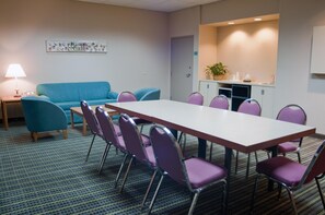 Uma sala de conferências com uma mesa comprida e cadeiras, uma pequena copa e um sofá azul, além de um abajur.