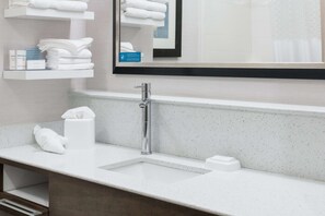 A modern bathroom with a white countertop, a sink, and a mirror above it. There are towels and toiletries on the countertop.