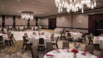 Un amplio salón de banquetes con mesas redondas dispuestas para cenar, con manteles blancos, sillas marrones y servilletas rojas.
