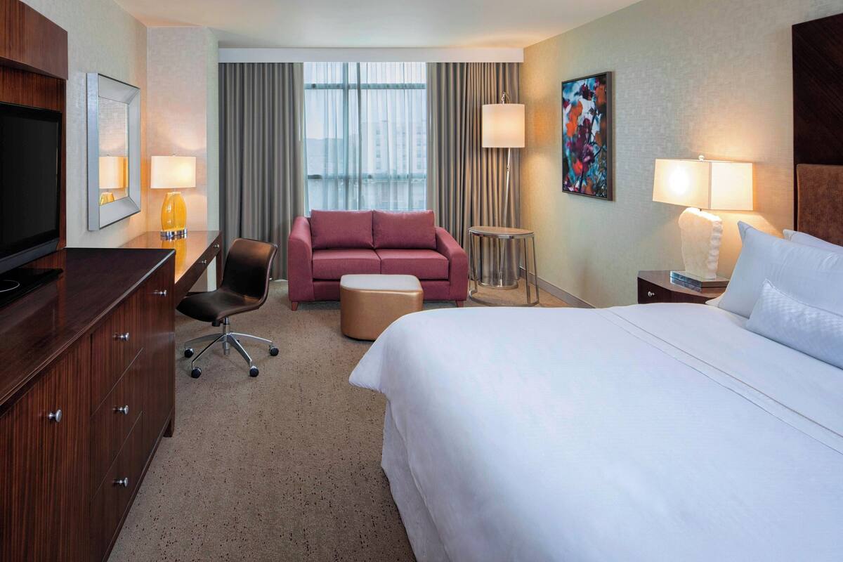 Une chambre d’hôtel avec un grand lit, un bureau avec une télévision, un canapé rose, un petit pouf, une chaise et une lampe.