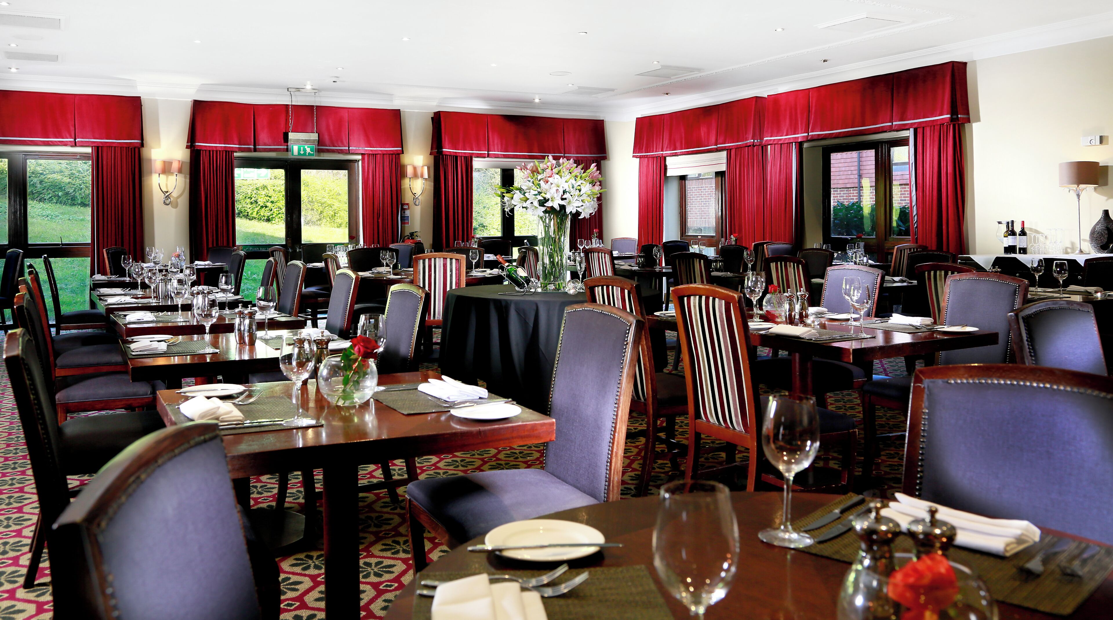 A formal dining room with multiple tables set for service, red drapery, and a view of greenery outside.