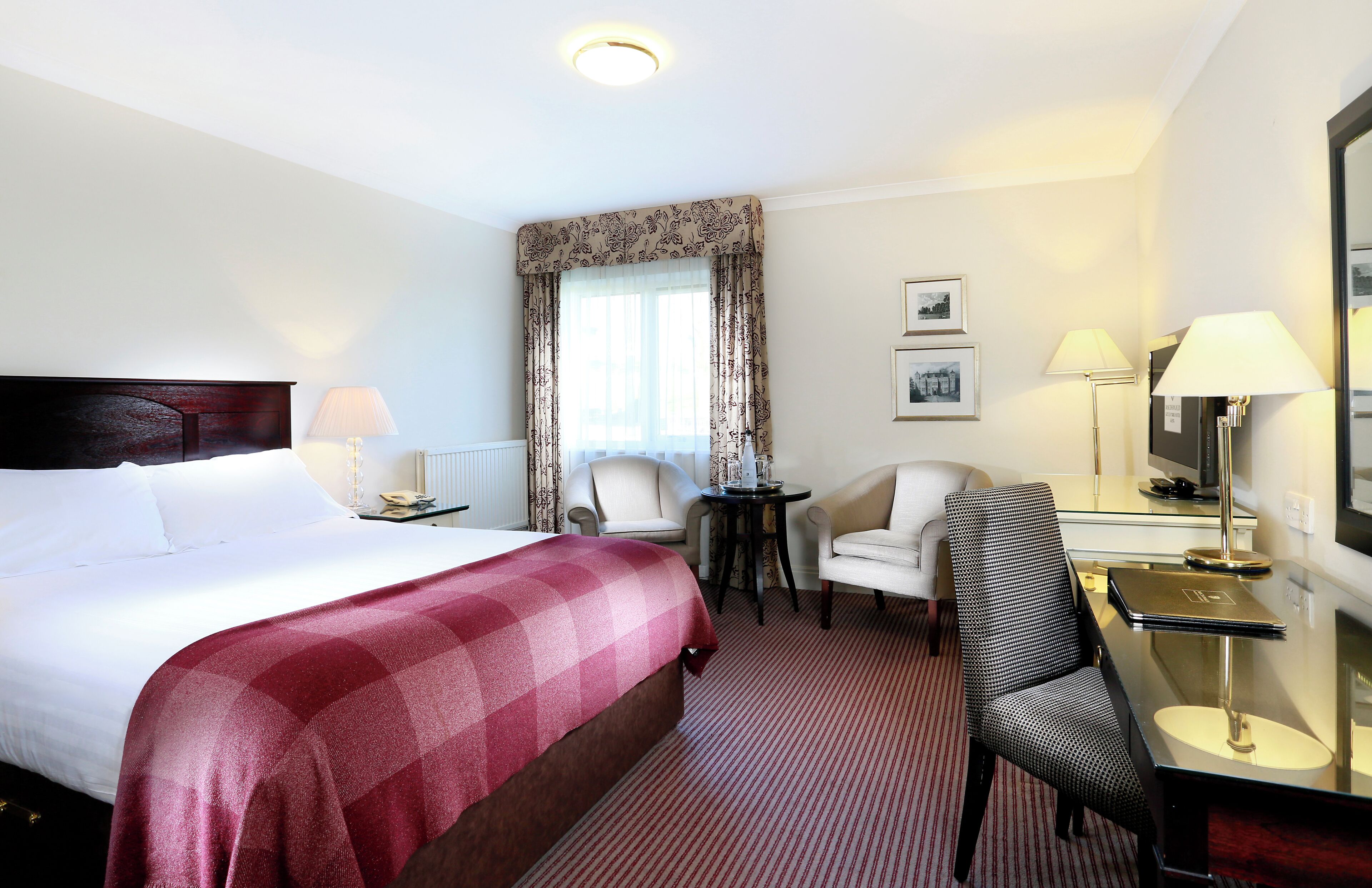 A hotel room with a large bed, a desk with a chair, a small table, and a lamp.