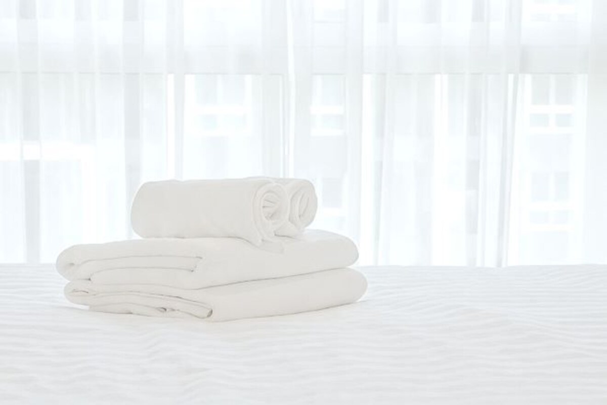 Una toalla blanca doblada cuidadosamente sobre una cama, con una cortina blanca translúcida al fondo.