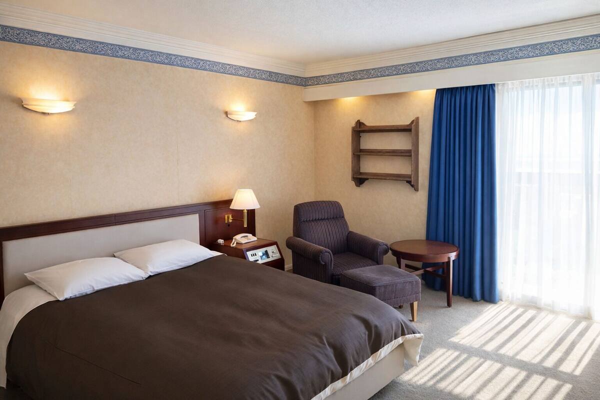 A hotel room with a large bed, a bedside table with a lamp, a chair, a small table, and a blue curtain.