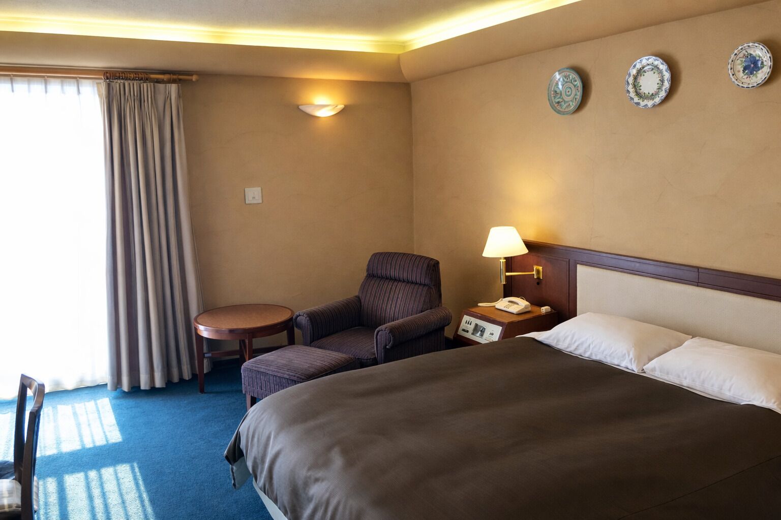 Habitación de hotel con una cama grande, una silla, una mesita, una lámpara y una mesita de noche con un teléfono.