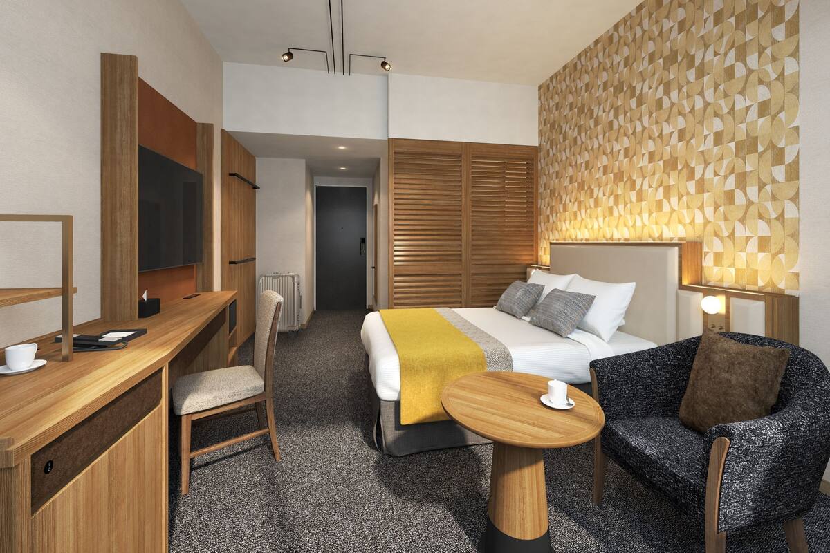 A hotel room with a bed, a desk, a chair, a small table, and a wall with a geometric pattern.