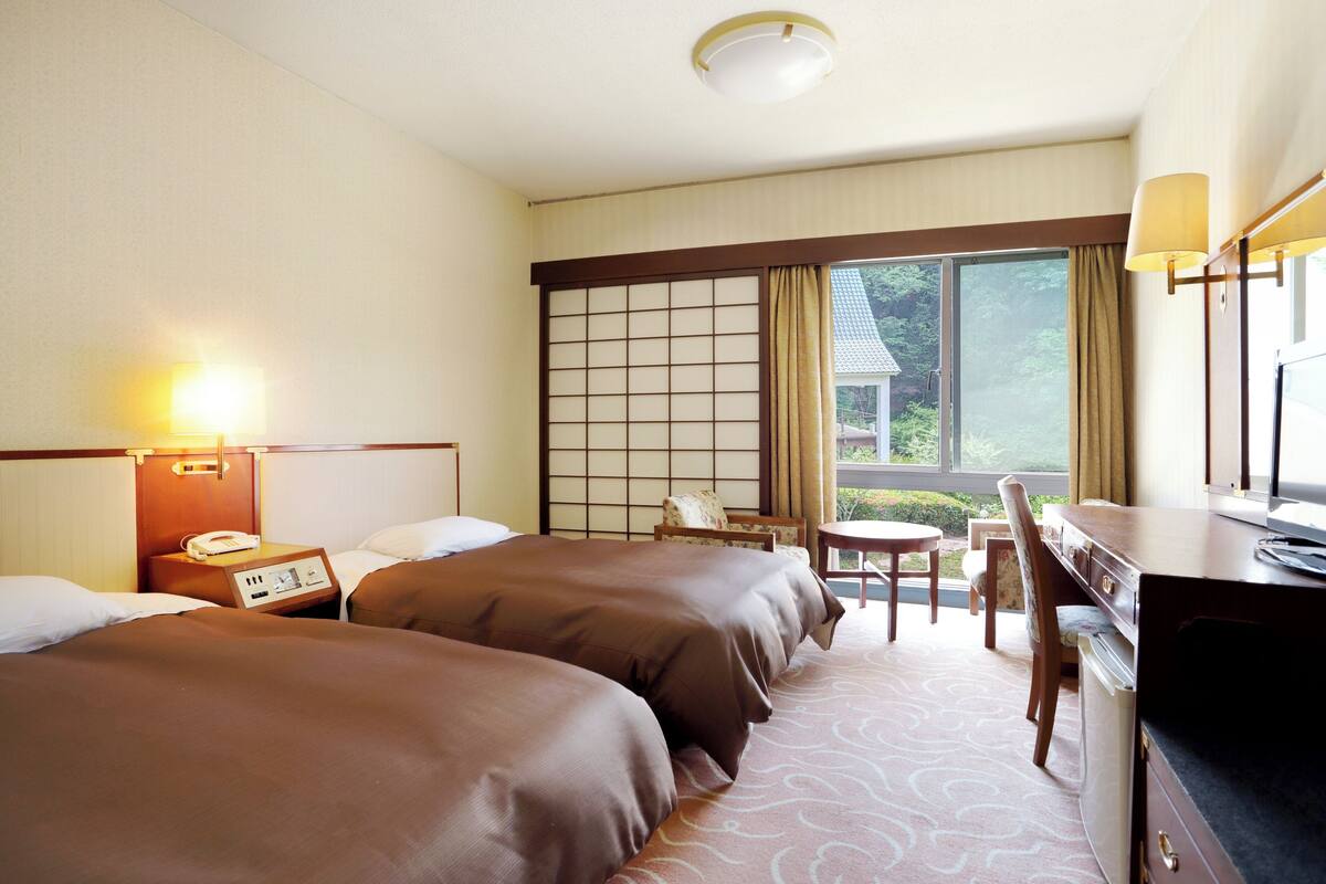 A hotel room with two beds, a desk, a chair, a small table, and a window with curtains.