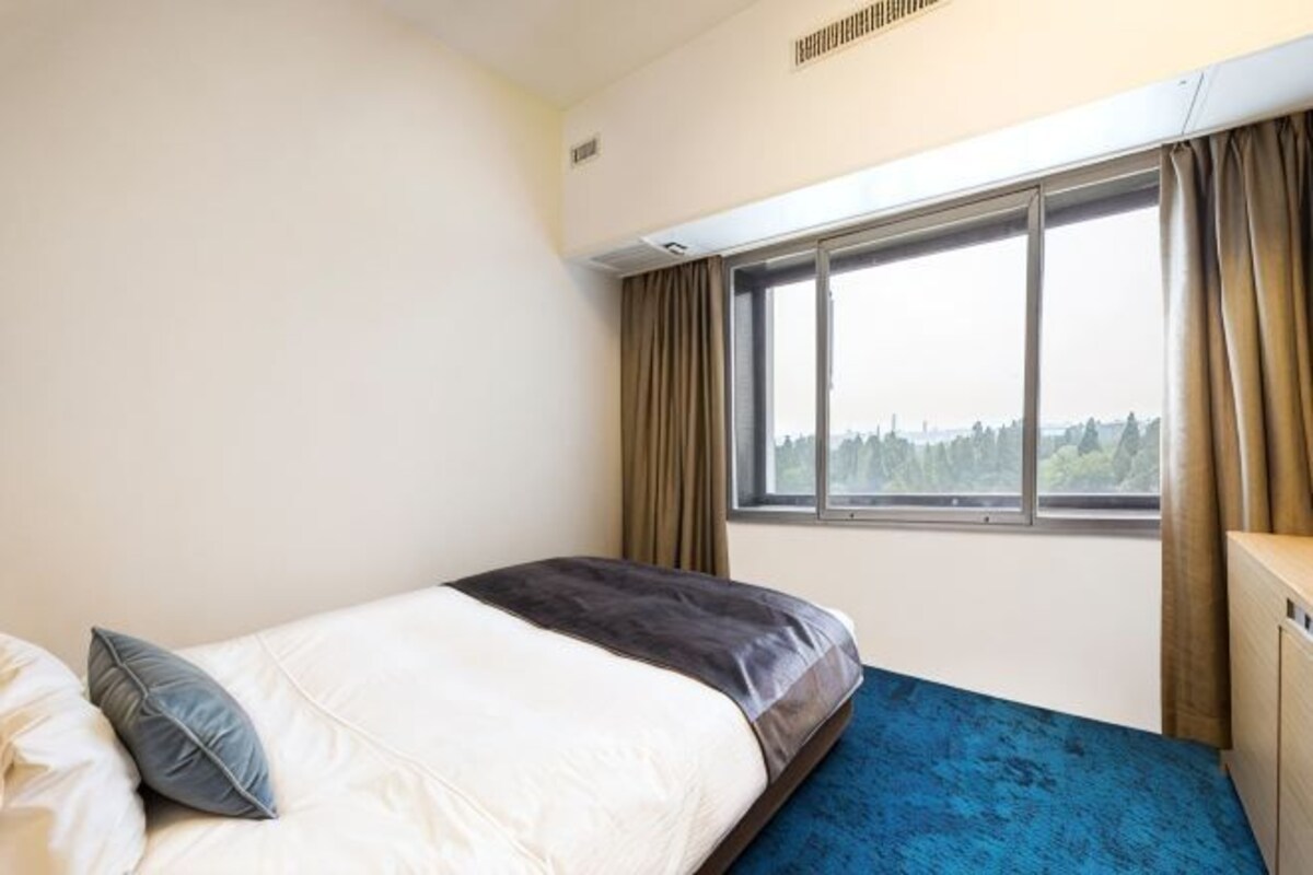Una cama individual con colchón azul, una ventana con cortinas y vistas a un paisaje.