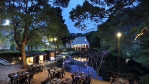 Un área de comedor al aire libre con mesas y sillas, rodeada de árboles y un edificio al fondo.