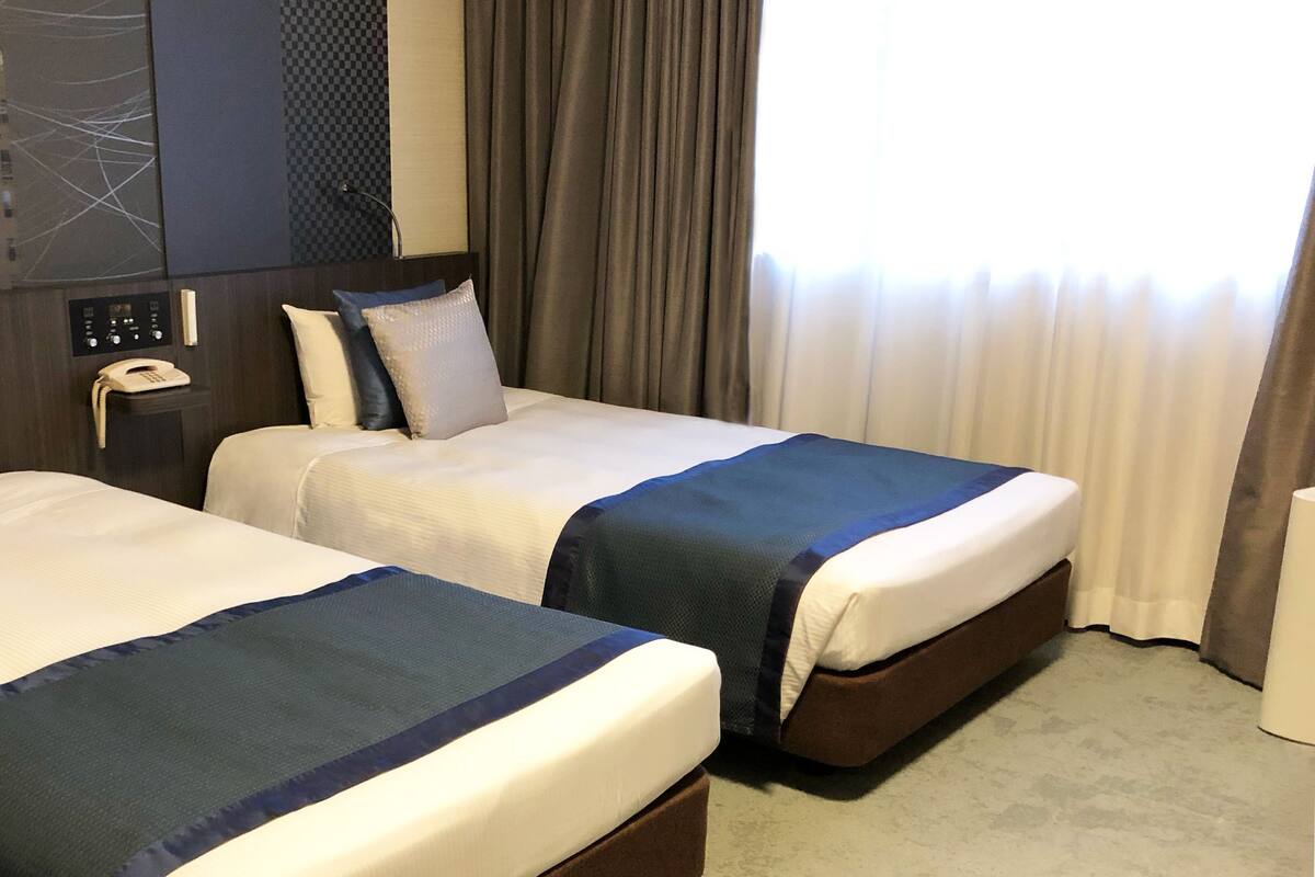 A hotel room with two beds, a control panel, a telephone, and a window with curtains.