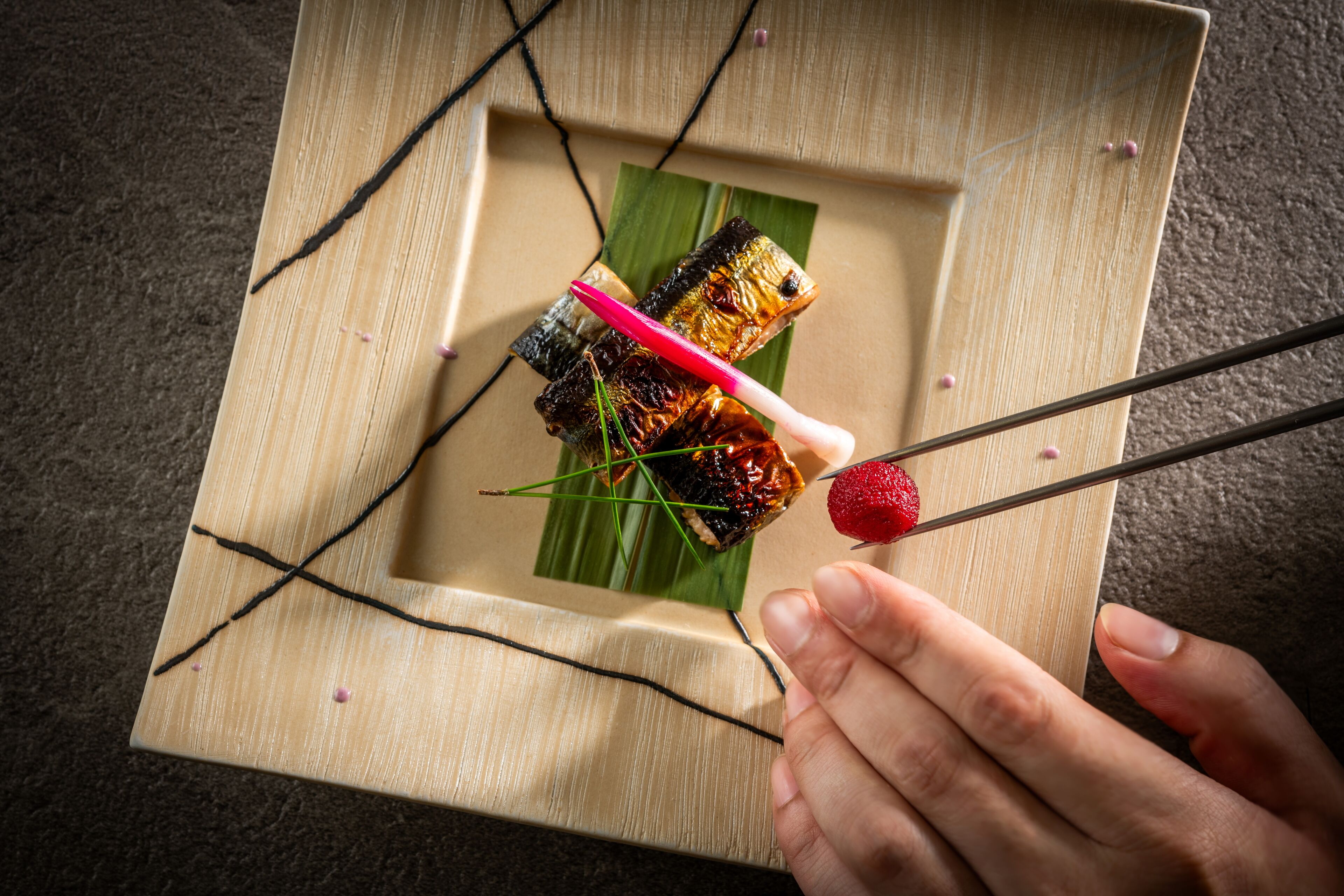 A wooden tray with a piece of food on a banana leaf, a hand holding chopsticks with a strawberry, and a small piece of food on a toothpick.