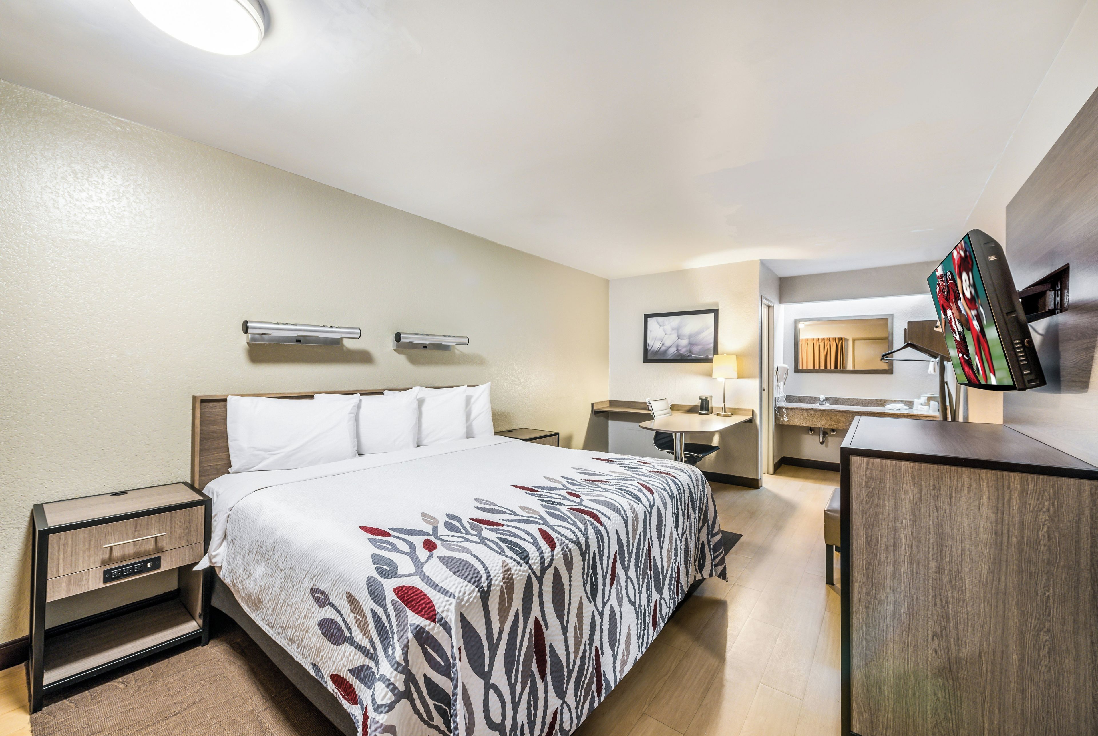 Une chambre d’hôtel avec un lit, des tables de chevet, un bureau, une chaise, une télévision et une salle de bain visible à travers une porte ouverte.