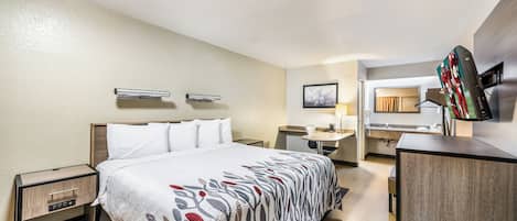 A hotel room with a bed, bedside tables, a desk, a chair, a TV, and a bathroom visible through an open door.