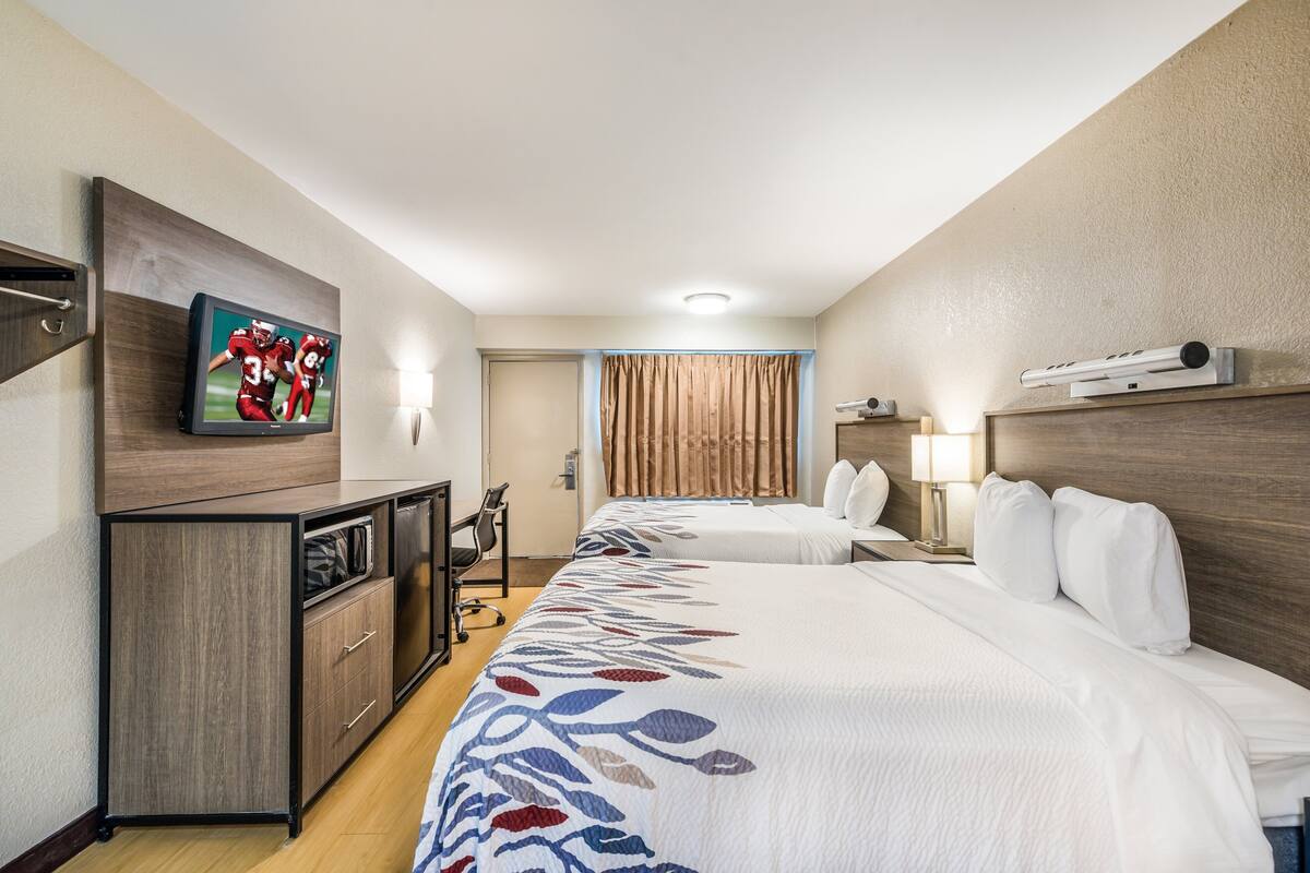 A hotel room with two beds, a TV mounted on the wall, a desk with a chair, and a window with curtains.