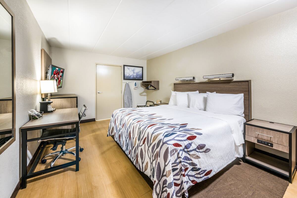 A hotel room with a bed, a desk, a chair, a nightstand, a lamp, a TV, and a door.