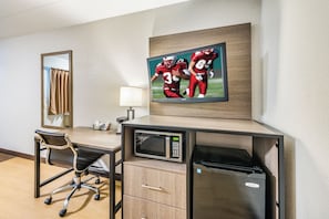Habitación de hotel con escritorio, una silla, microondas, refrigerador, televisor montado en la pared y espejo.