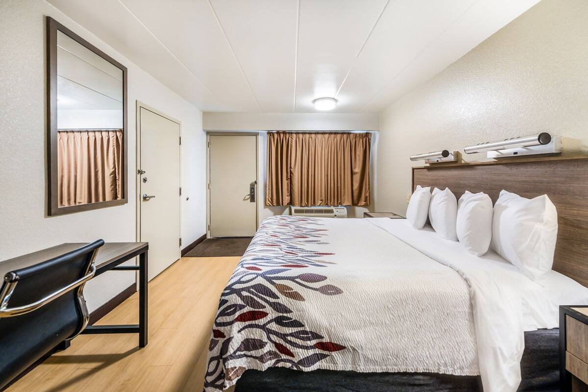 A hotel room with a bed, a desk, a chair, a mirror, and a window with curtains.